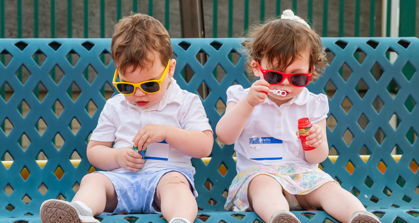 Two young children wearing sunglasses sit together on a bench; one blows bubbles while the other examines a bubble wand.