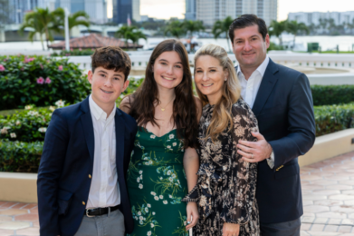 A family of four, dressed in formal attire, poses together outside.