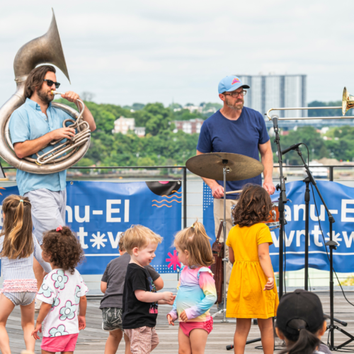 Three musicians play on an outdoor stage at Temple Emanu-El as children dance and enjoy the sunny day in front of them.