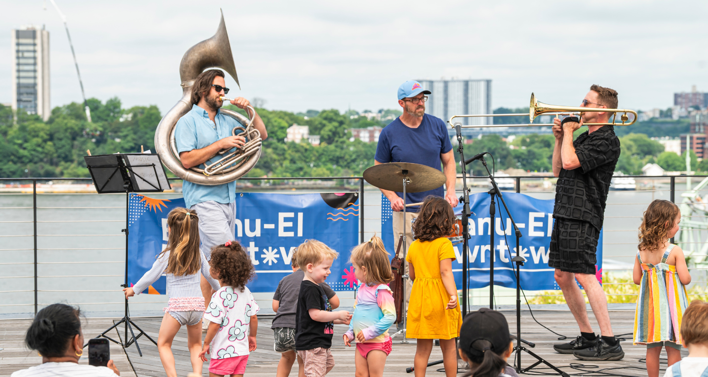 Three musicians play on an outdoor stage at Temple Emanu-El as children dance and enjoy the sunny day in front of them.