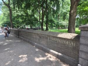 People walk beside a stone wall labeled Artisans Gate in a tree-lined area near Temple Emanu-El in New York City on a sunny day.