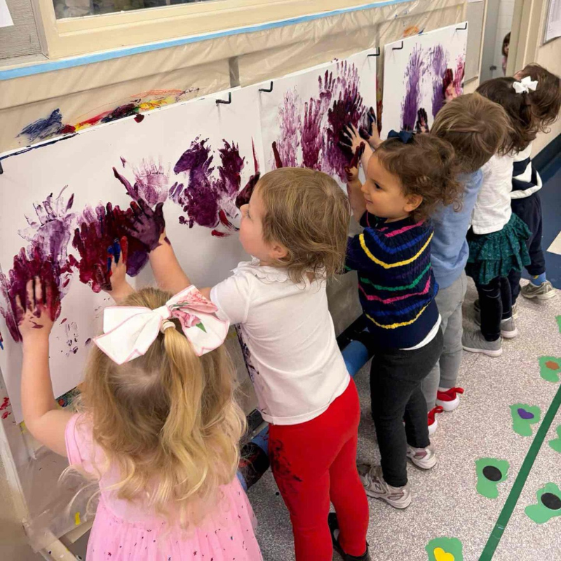 Five young children joyfully use their hands to paint on large sheets of paper taped to a classroom wall at Temple Emanu-El.