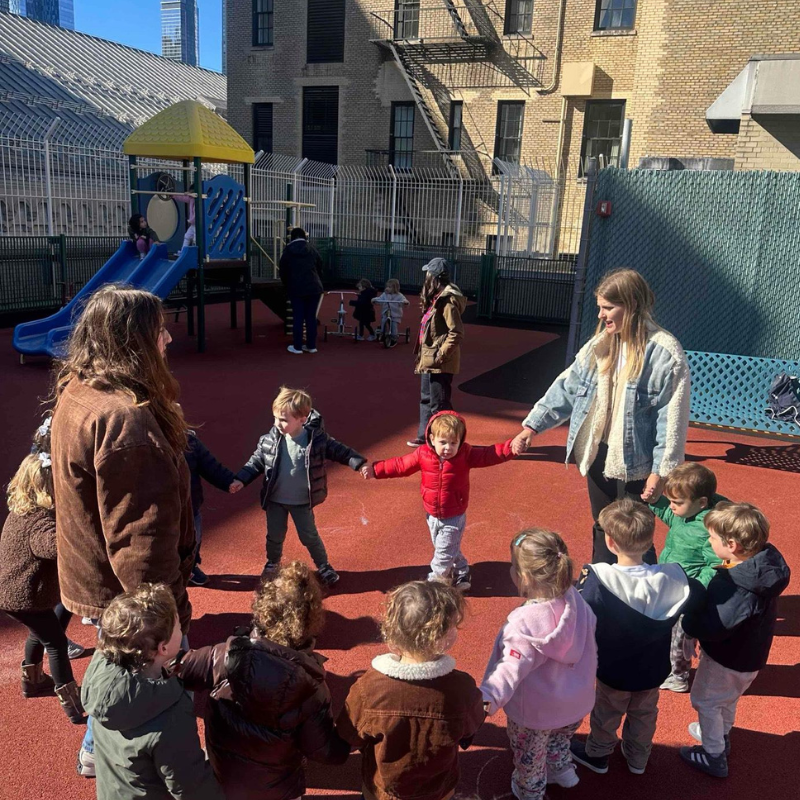 Children and two adults join hands in a circle on the Temple Emanu-El playground, with a slide visible in the background.