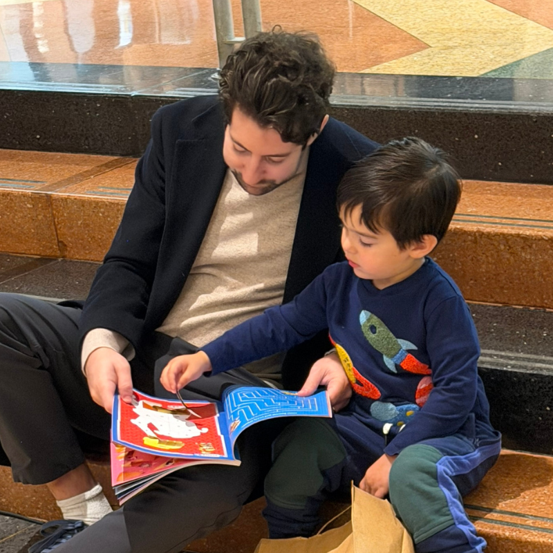 An adult and a child sit on steps at Temple Emanu-El, reading a colorful book together, both focused on the pages.