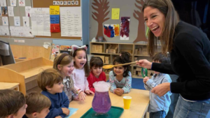 A teacher and young children joyfully observe a bubbling purple liquid experiment together in a Temple Emanu-El classroom.