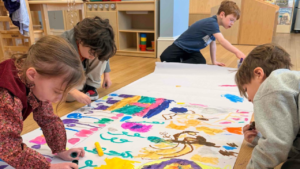Four young children draw and color together on a large sheet of paper spread out on the classroom floor at Temple Emanu-El.