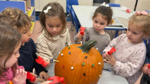 Five young children use toy hammers to gently tap colorful pegs into a large pumpkin during a festive activity at Temple Emanu-El.