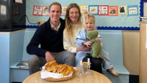 A joyful family gathers in a classroom at Temple Emanu-El, sharing pastries and a candle, as a child holds a green toy dinosaur.
