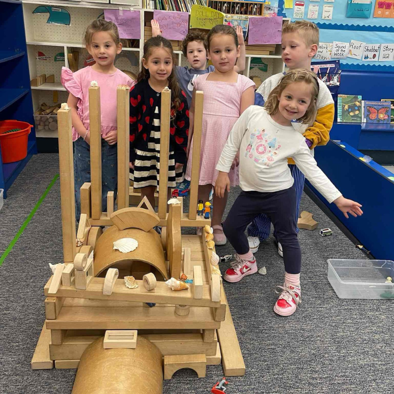 Six young children smile behind a large wooden block castle they built together in a Temple Emanu-El classroom.