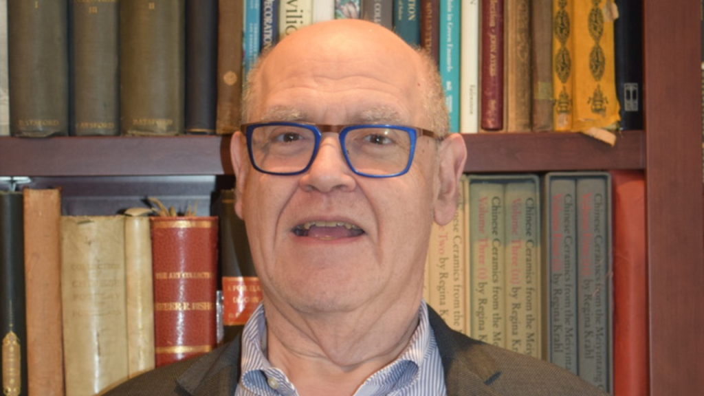 Elderly man with glasses smiling warmly in front of a bookshelf at Temple Emanu-El, surrounded by books.