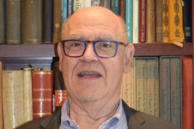 Elderly man with glasses smiling warmly in front of a bookshelf at Temple Emanu-El, surrounded by books.