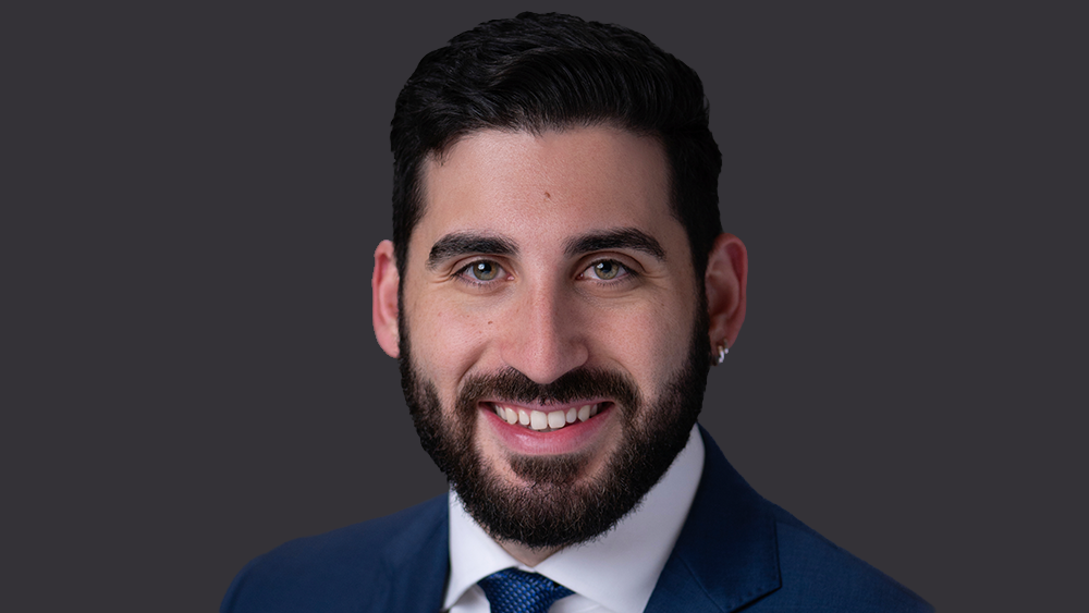 Smiling man with dark hair and beard, dressed in a blue suit and tie, posed against a dark gray background.