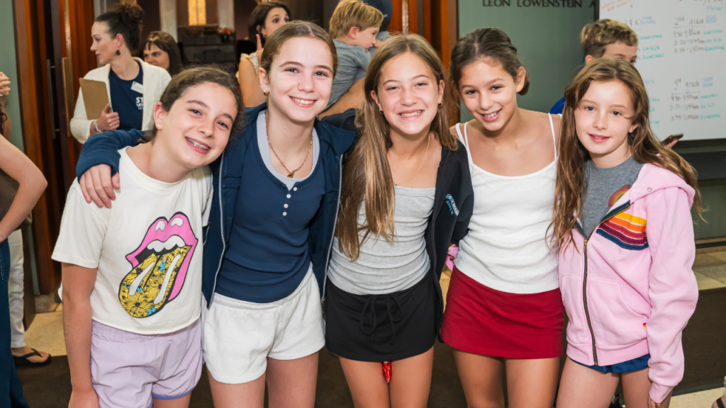 Five smiling girls stand arm-in-arm inside Temple Emanu-El, joyfully posing together for a group photo.