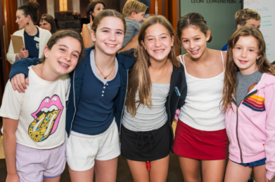 Five smiling girls stand arm-in-arm inside Temple Emanu-El, joyfully posing together for a group photo.