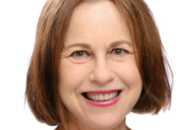 Smiling woman with straight brown hair wearing a modest grey top, photographed against a plain white background.