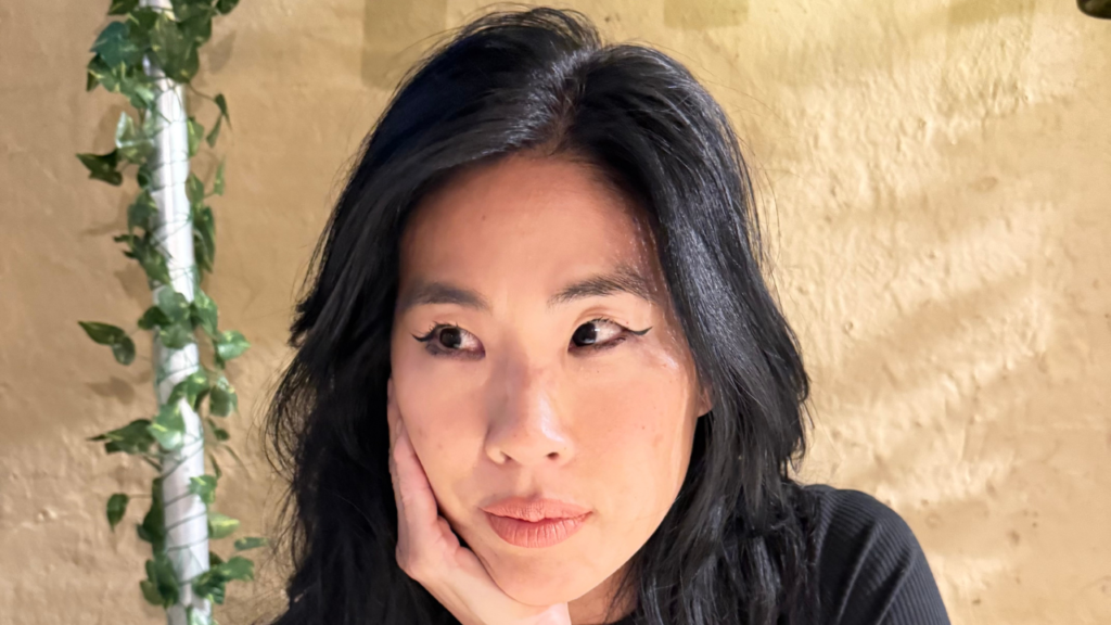 A woman with long black hair rests her chin on her hand, appearing thoughtful, seated against a neutral beige background.