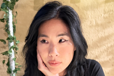 A woman with long black hair rests her chin on her hand, appearing thoughtful, seated against a neutral beige background.