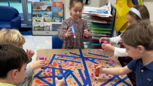 Five young children create colorful art together using paint daubers and paper strips in a classroom at Temple Emanu-El.