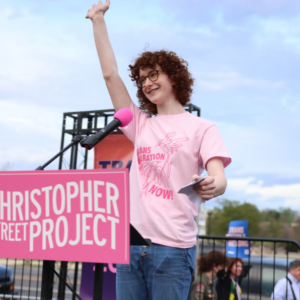 A person in a pink shirt speaks at a podium labeled CHRISTOPHER STREET PROJECT.