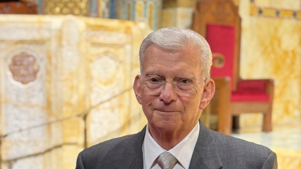 An older man in a gray suit and tie stands in a beautifully lit sanctuary adorned with gold and red accents at Temple Emanu-El.