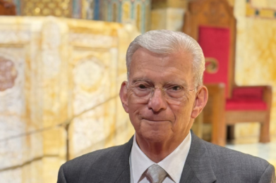 An older man in a gray suit and tie stands in a beautifully lit sanctuary adorned with gold and red accents at Temple Emanu-El.