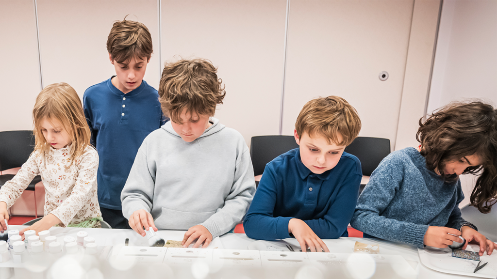 Five children gather at a table at Emanu-El Downtown, engaged in arts and crafts activities using a variety of colorful supplies.