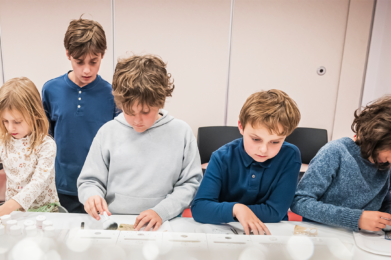 Five children gather at a table at Emanu-El Downtown, engaged in arts and crafts activities using a variety of colorful supplies.