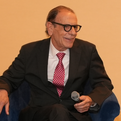 Distinguished older man in a suit and glasses smiles while holding a microphone at Temple Emanu-El, with water on a table nearby.