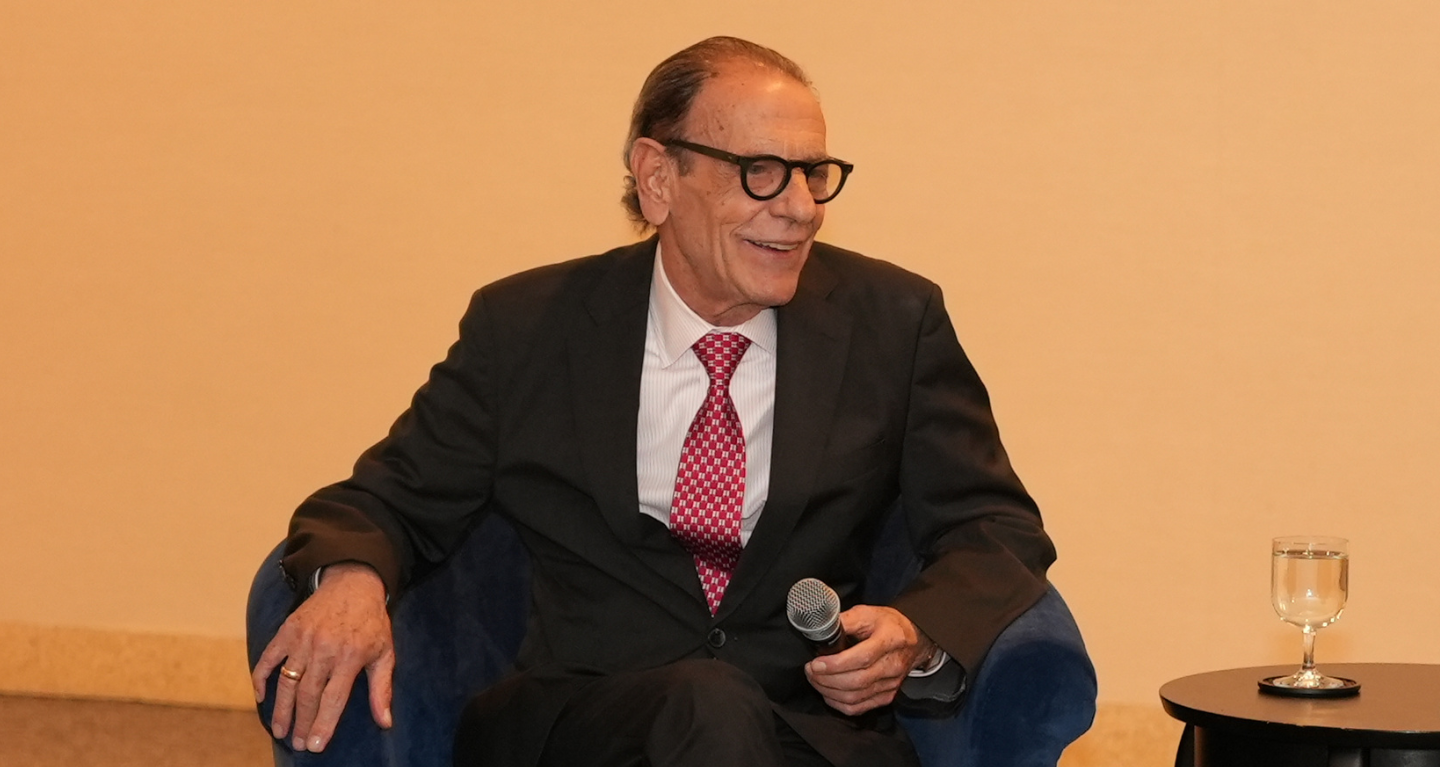 Distinguished older man in a suit and glasses smiles while holding a microphone at Temple Emanu-El, with water on a table nearby.