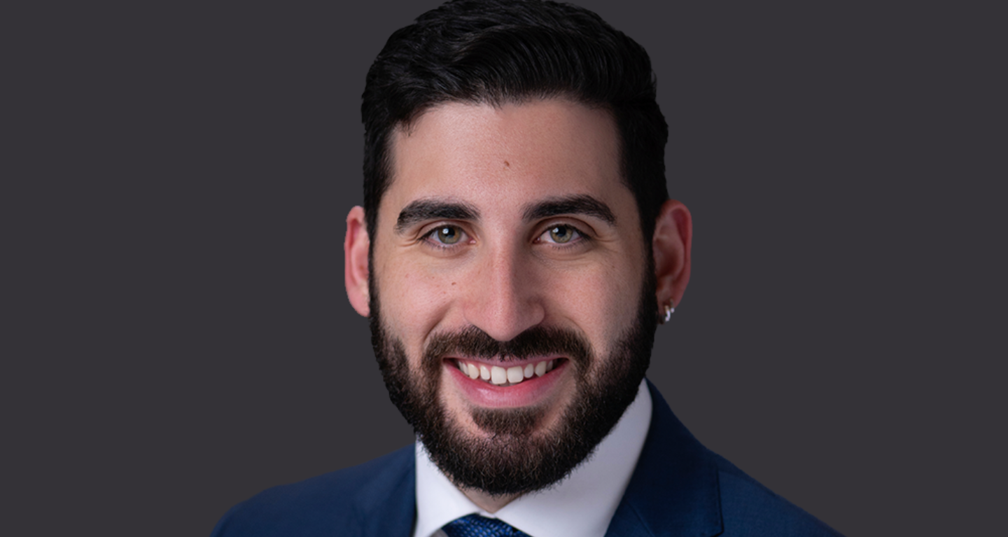 Man with short dark hair and beard, dressed in a blue suit and tie, smiling warmly against a dark background.