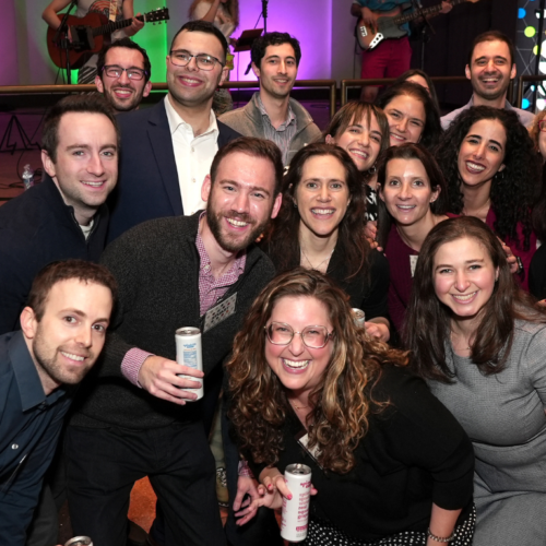 A group of smiling adults gathers together indoors at a Temple Emanu-El event, with colorful lights illuminating the background.