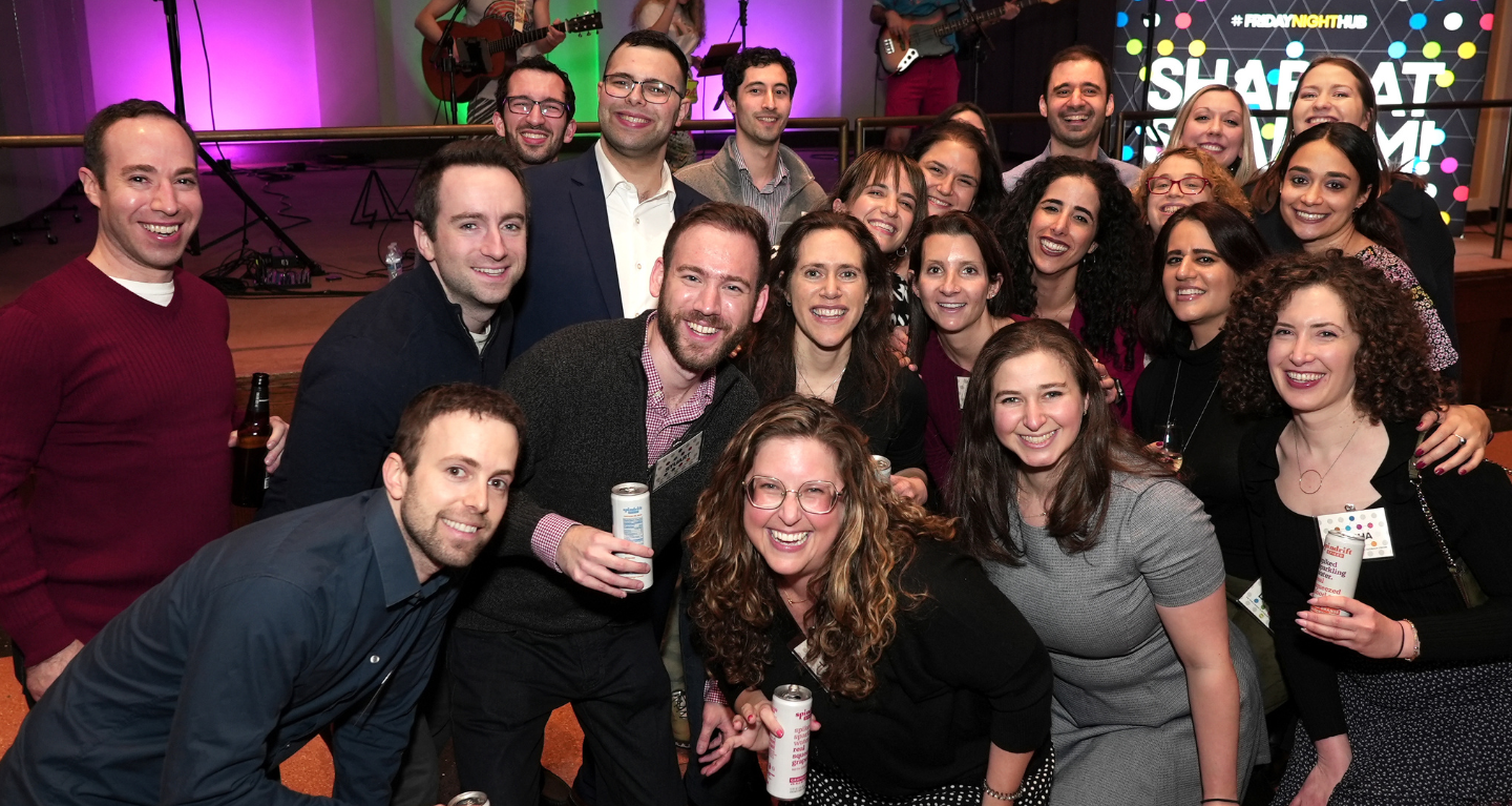 A group of smiling adults gathers together indoors at a Temple Emanu-El event, with colorful lights illuminating the background.