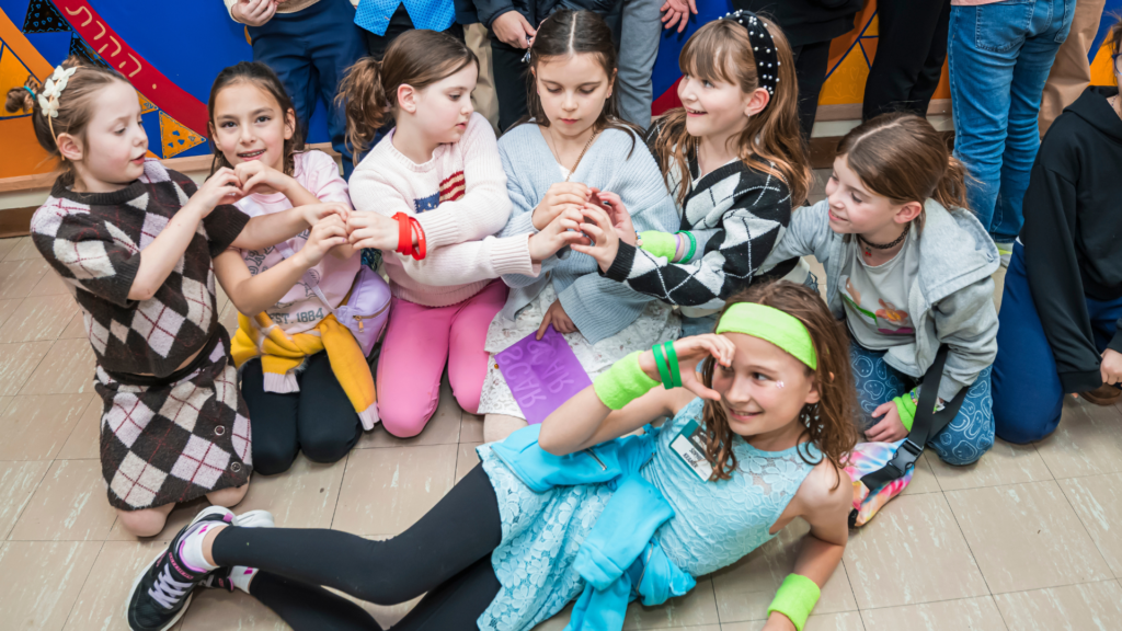 Seven children gather indoors at Temple Emanu-El, some sitting and some kneeling, many forming heart shapes with their hands.