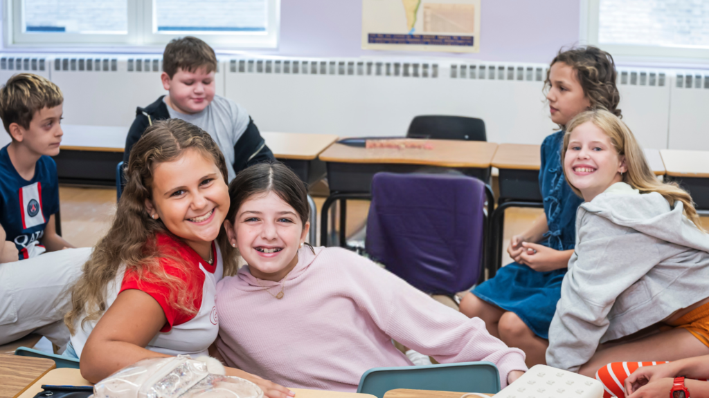 Six children sit and smile together in a bright classroom at Temple Emanu-El, with desks and large windows in the background.