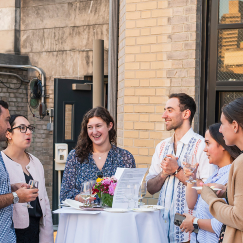 Five people stand around a tall table outdoors on a rooftop at Temple Emanu-El, enjoying conversation and refreshments during a community gathering.
