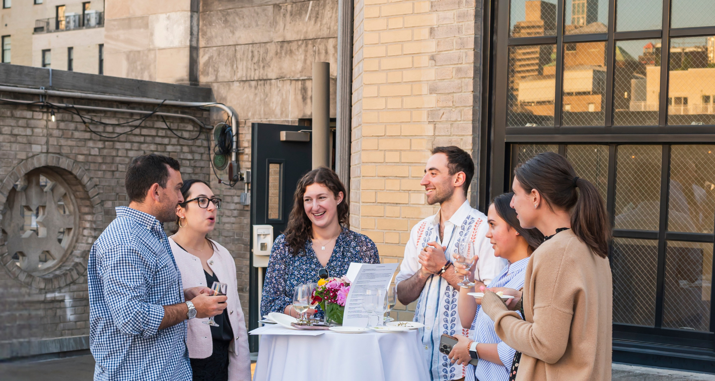 Five people stand around a tall table outdoors on a rooftop at Temple Emanu-El, enjoying conversation and refreshments during a community gathering.