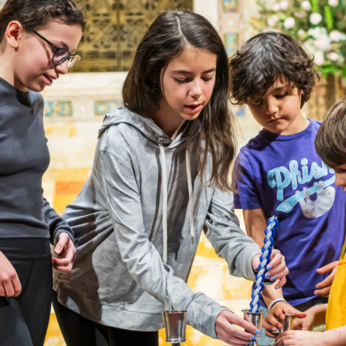 Four children participate in lighting a Havdalah candle together indoors at Temple Emanu-El, with flowers and chairs visible in the background.