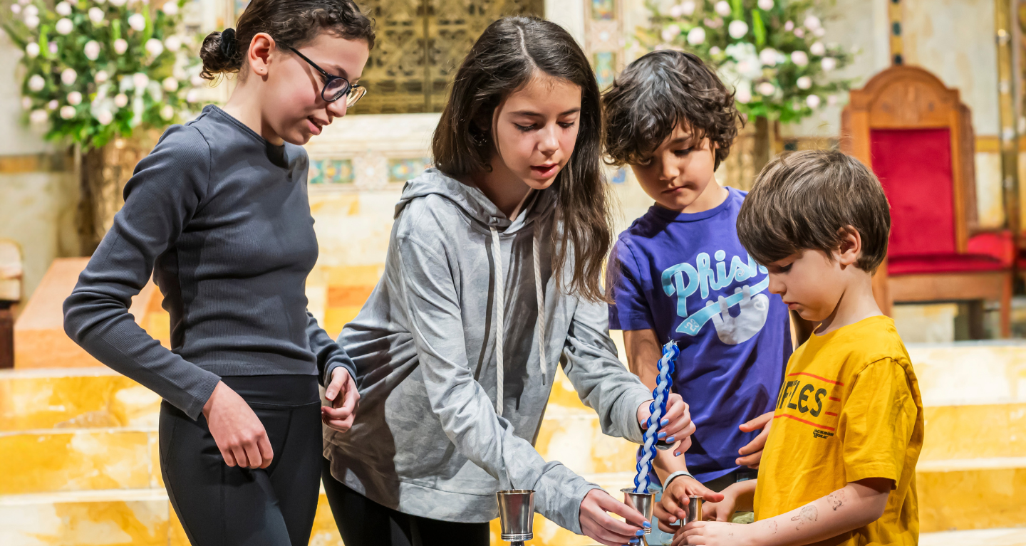 Four children participate in lighting a Havdalah candle together indoors at Temple Emanu-El, with flowers and chairs visible in the background.