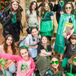Group of girls in green outfits smiling and posing together indoors at a Temple Emanu-El event, some wearing heart-shaped glasses and holding festive props.