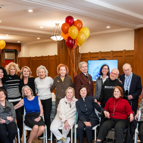 A group of adults gather indoors at Temple Emanu-El, smiling at the camera with festive balloons in the background.