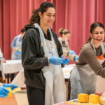Three women wearing gloves and aprons smile as they prepare sandwiches together during a Temple Emanu-El community service event.