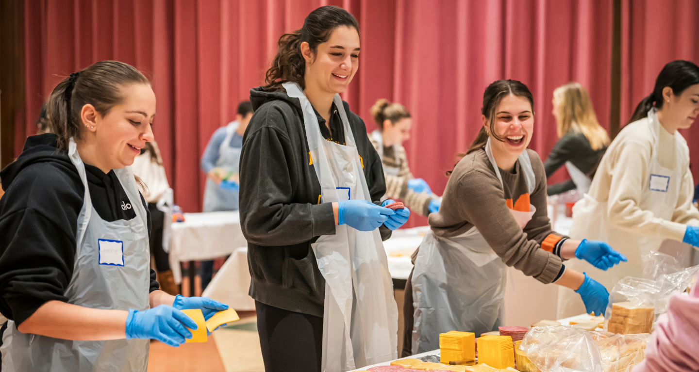 Three women wearing gloves and aprons smile as they prepare sandwiches together during a Temple Emanu-El community service event.