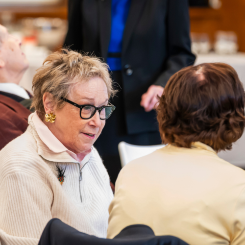 Elderly women engaged in conversation at a table during a formal gathering at Temple Emanu-El, with refreshments and attendees nearby.