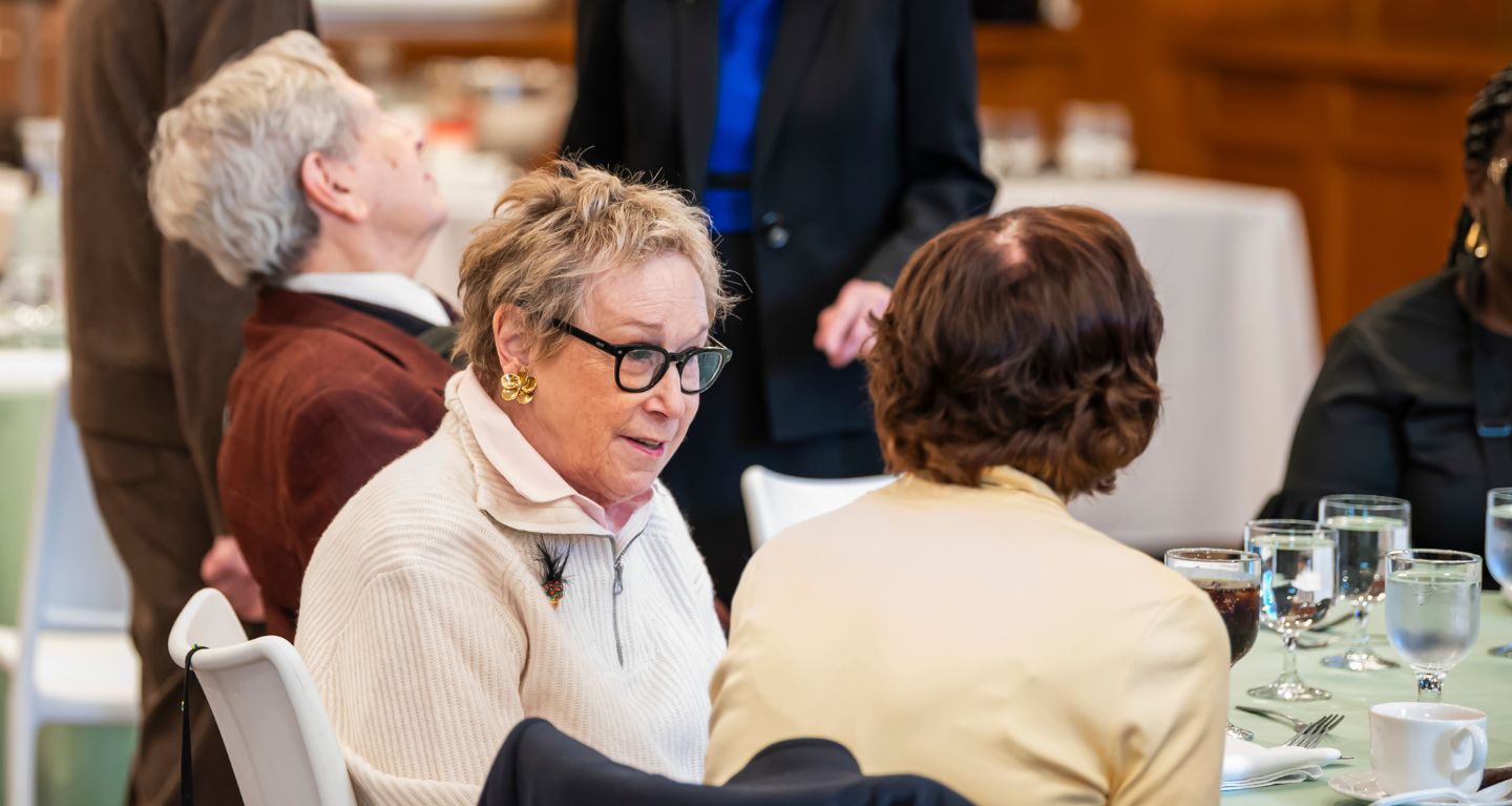 Elderly women engaged in conversation at a table during a formal gathering at Temple Emanu-El, with refreshments and attendees nearby.