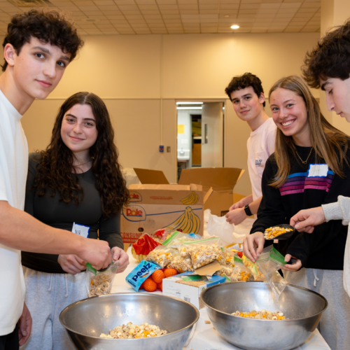 Five teens smile as they prepare snacks together at a table in a brightly lit room at Temple Emanu-El.