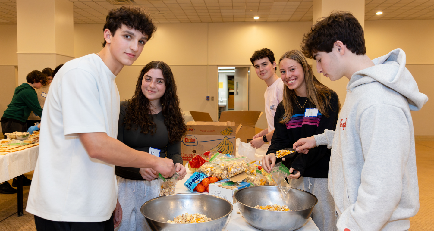 Five teens smile as they prepare snacks together at a table in a brightly lit room at Temple Emanu-El.