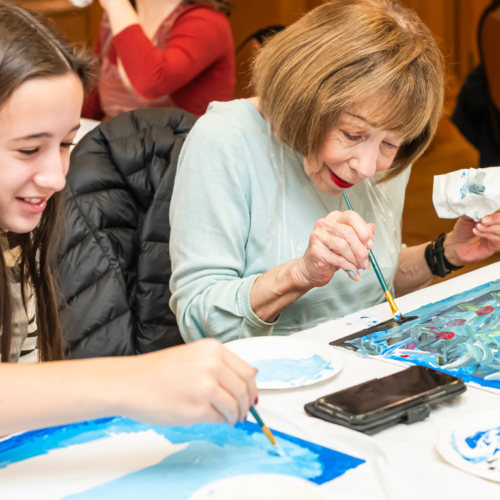 An older woman and a girl joyfully paint together at a table filled with art supplies and vibrant artwork during an intergenerational program at Temple Emanu-El.
