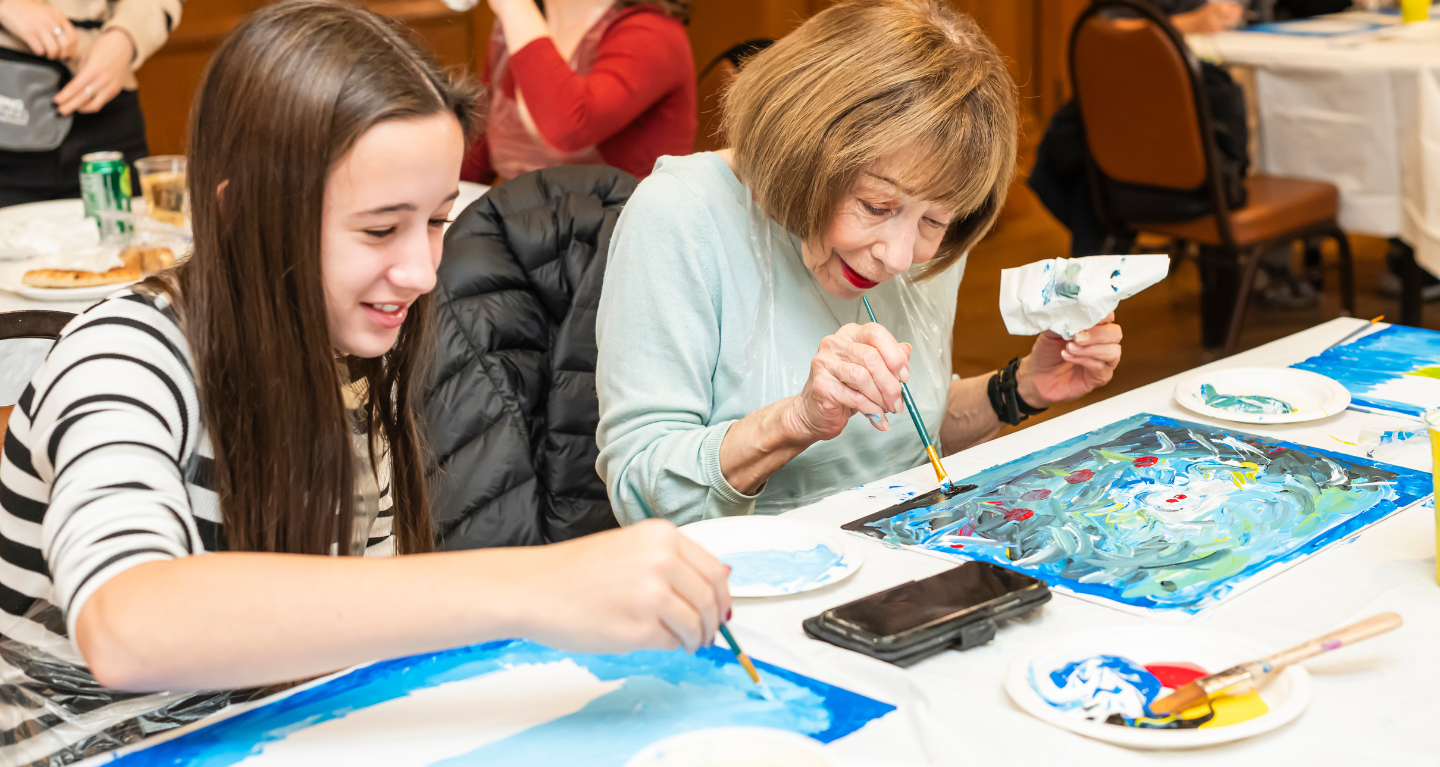 An older woman and a girl joyfully paint together at a table filled with art supplies and vibrant artwork during an intergenerational program at Temple Emanu-El.