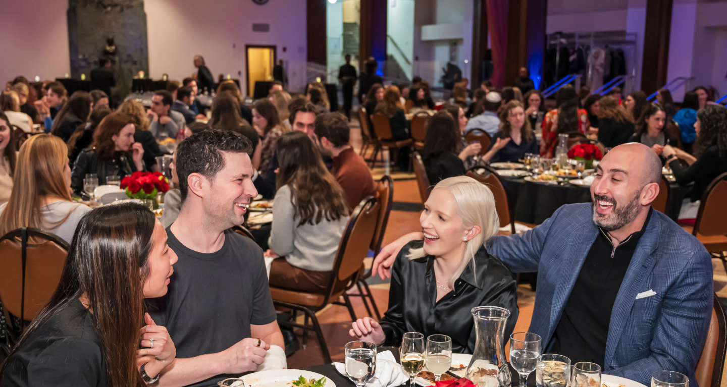 Four people smiling and sharing laughter around a dinner table in a beautifully decorated, bustling event space at Temple Emanu-El.