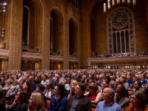 A large congregation gathers in Temple Emanu-El's majestic sanctuary with tall windows, attentively observing a service.